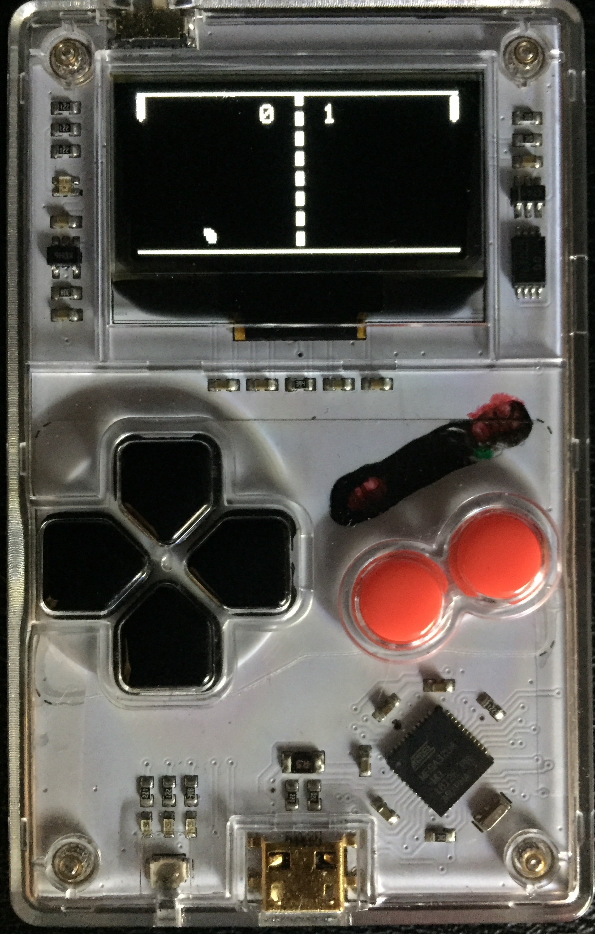 Photograph of the clear-shell Arduboy, board and chip visible through the case, running the game: a paddle at each side of the screen, a dashed centre line, the ball mid-flight and the score reading 0 to 1.