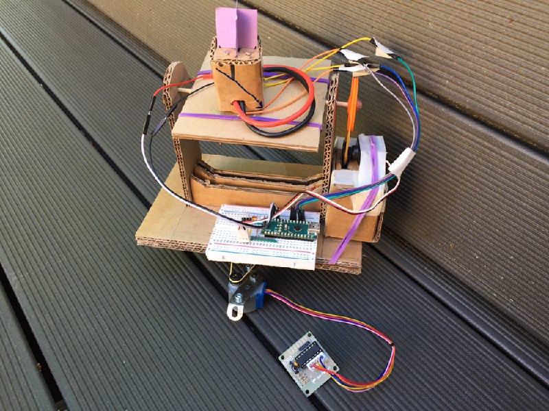 The assembled prototype outdoors on grey decking: a two-level corrugated cardboard frame carrying both drives, the sensor head with its purple vane on the upper tilting flap, a small Arduino-compatible board on a breadboard on the lower deck, coloured jumper wires running between the two levels, a servo with an orange horn on the right, and a small green breakout board at the end of a four-wire lead.