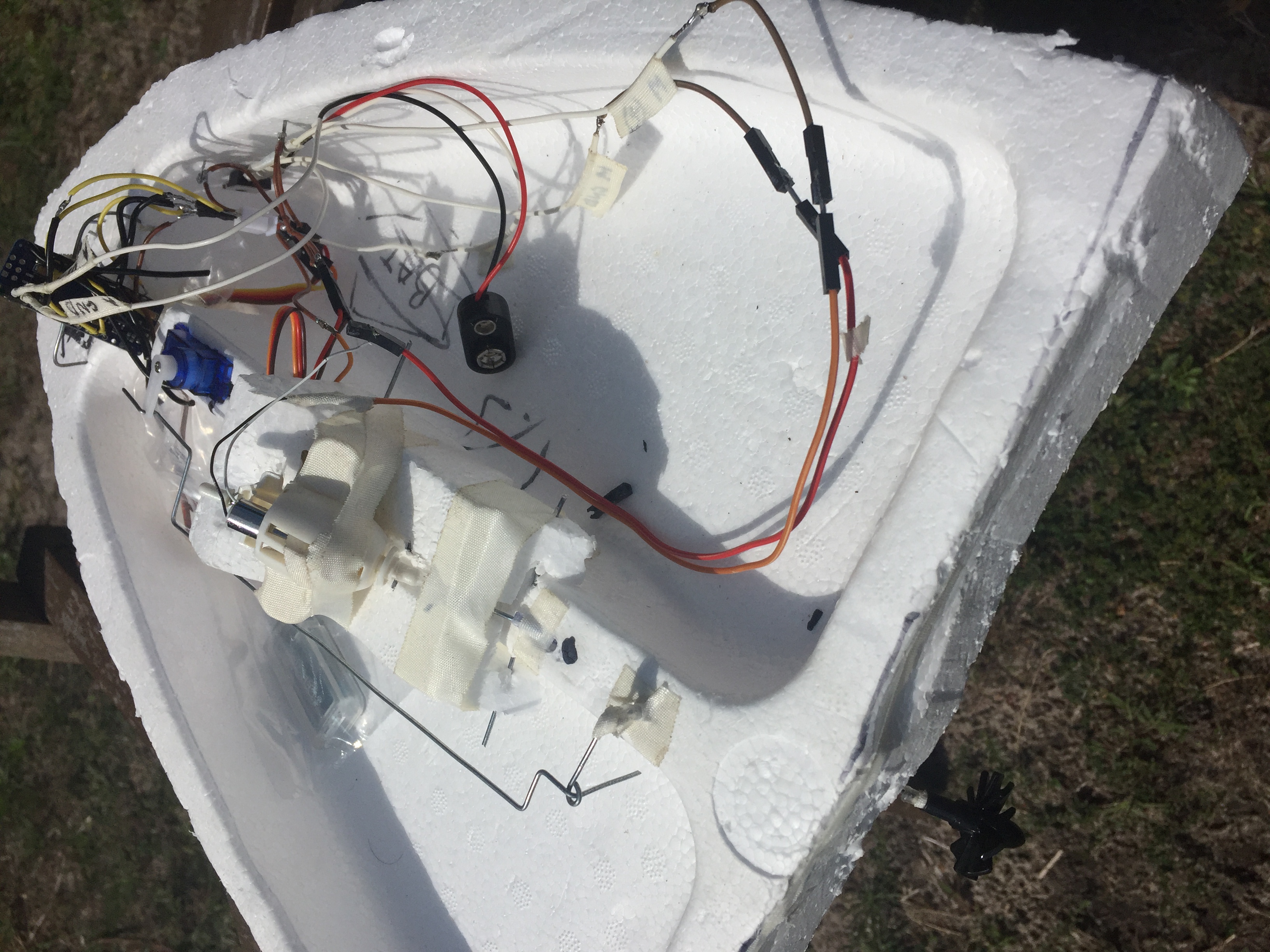 Overhead photograph of the finished boat sitting on grass: a hand-cut white polystyrene hull holding a small blue servo, a DC motor strapped down with white cloth tape, a bent steel pushrod running along the hull, a small blue radio module and a loom of jumper wires, with the word BAT written on the foam in pencil and a black propeller at one end of the hull.