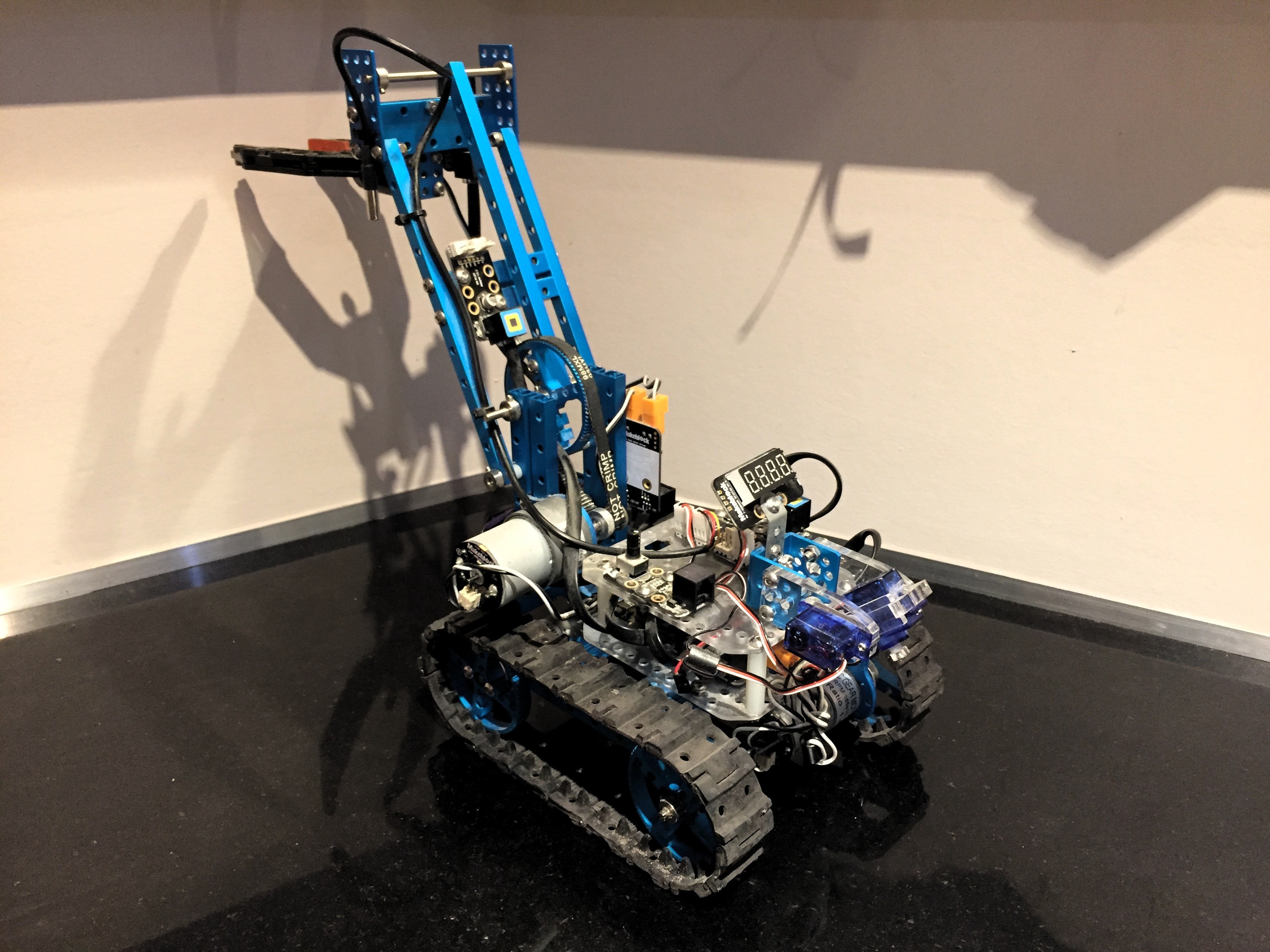 Photograph of the first robot on a dark floor: a blue aluminium Makeblock chassis on rubber tracks, a tall boom arm raised at an angle with a black gripper at its top end, a geared DC motor, small purple servos, a four-digit seven-segment display and a bundle of wiring across the deck.