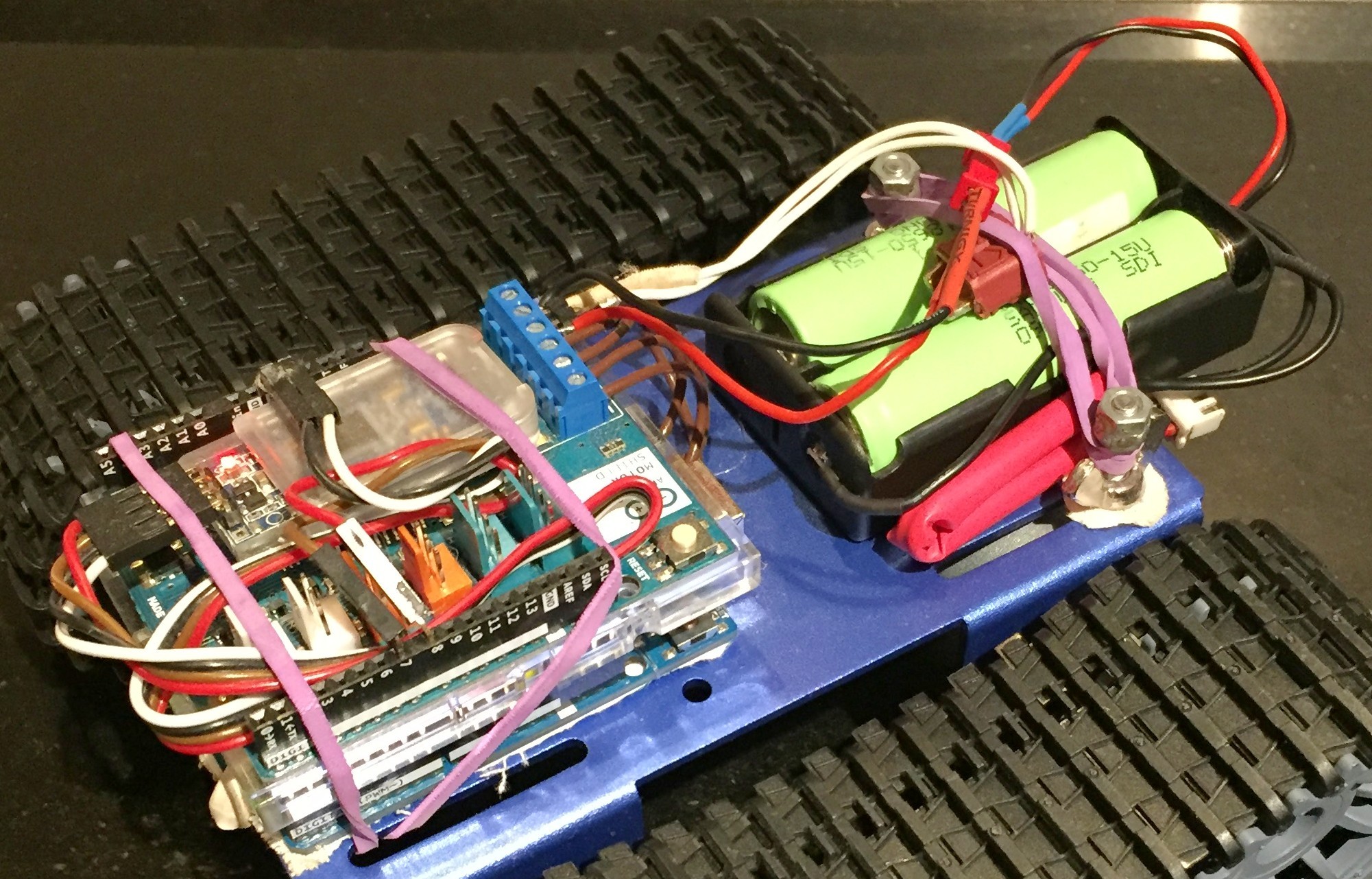 Close-up photograph of the second robot's deck: a stack of Arduino boards including a motor shield with a blue screw terminal block, a small module in a clear plastic case, all strapped down with purple rubber bands, next to a black holder of four green lithium-ion cells wired to a red connector, mounted on a blue aluminium chassis between two rubber tracks.