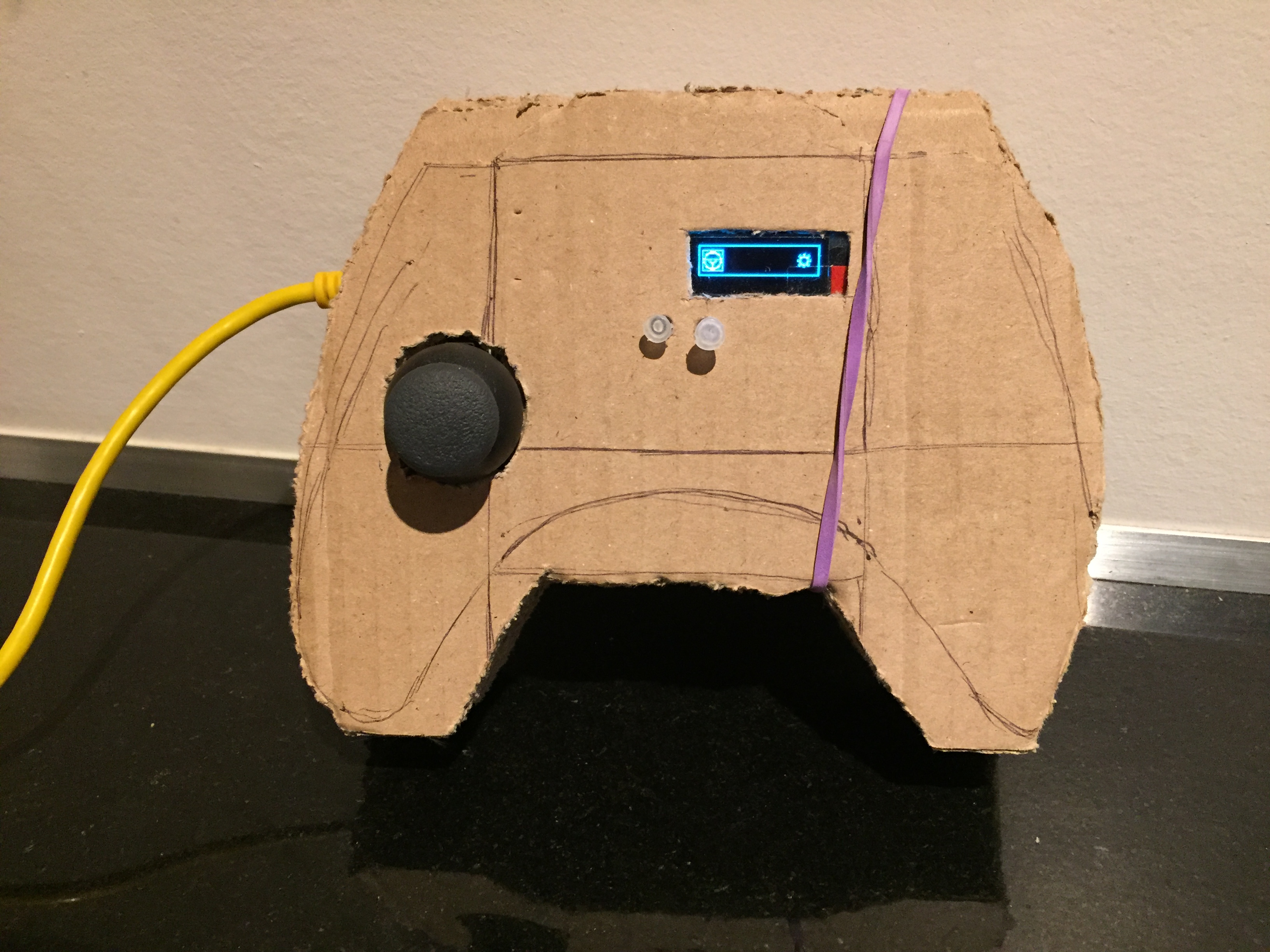 Photograph of the hand-made transmitter: a cardboard shell cut to the outline of a game controller with the design still drawn on it in pencil, a black thumb joystick pushed through a hole on the left, two small white buttons in the middle, a lit blue OLED screen showing two icons, and a purple rubber band stretched over the case.